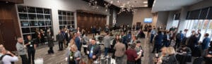 A large group of people gathers in a modern room for a corporate event. They are standing, conversing, and drinking beverages. An engaging emcee keeps the atmosphere lively. The room features high ceilings, large windows, and contemporary decor.